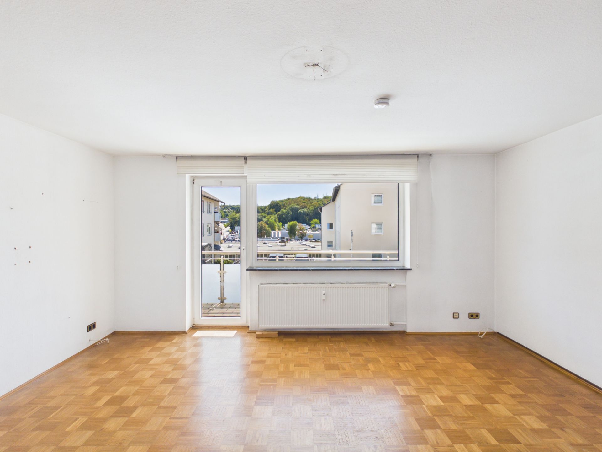 Leeres Zimmer mit Parkettboden, großem Fenster, Balkontür und Blick nach draußen.