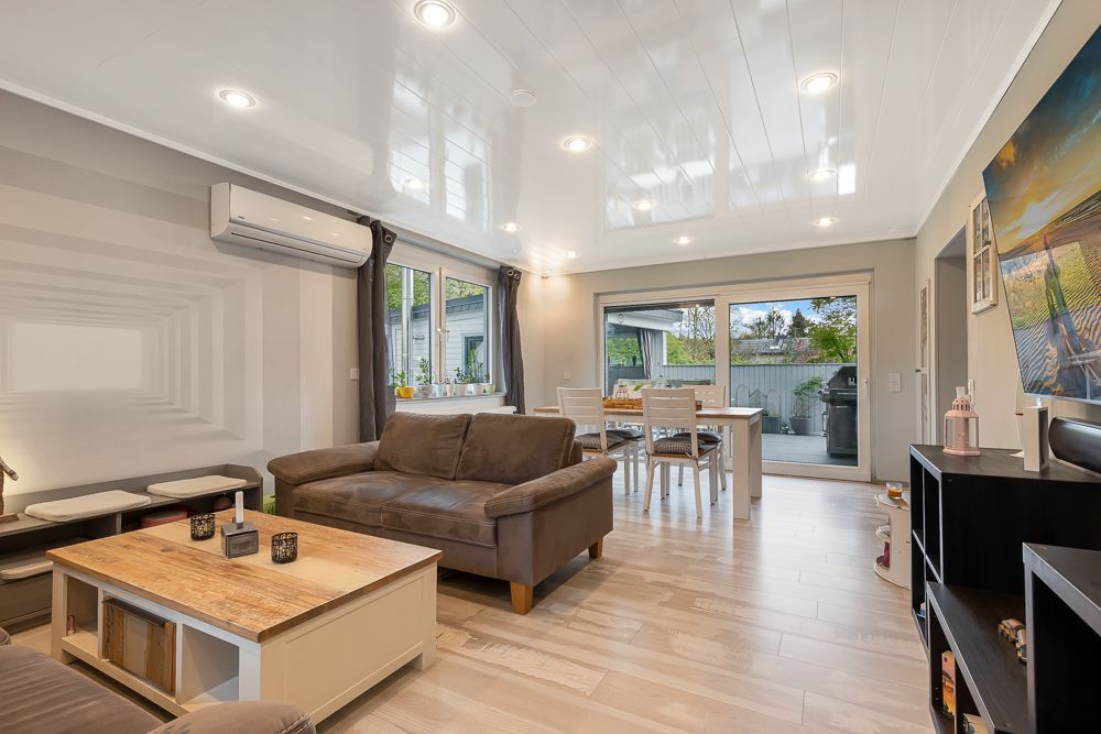 Helles Wohnzimmer mit braunem Sofa, Esstisch, großem Fenster und Zugang zur Terrasse.