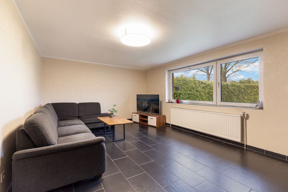Wohnzimmer mit grauem Ecksofa, Couchtisch, Fernseher und großem Fenster mit Blick ins Grüne.