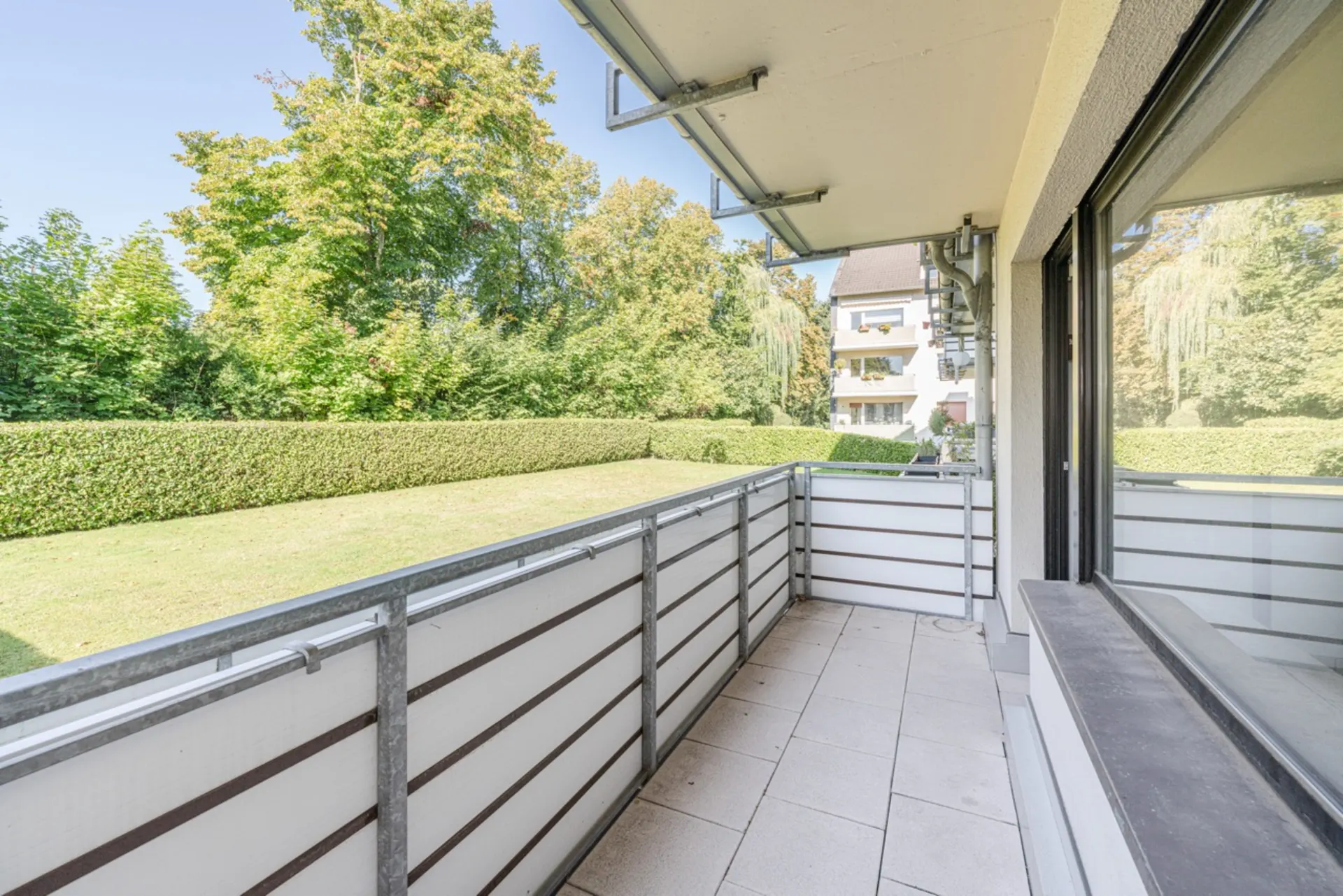 Balkon mit Metallgeländer und Blick auf einen grünen Garten mit Büschen und Bäumen.
