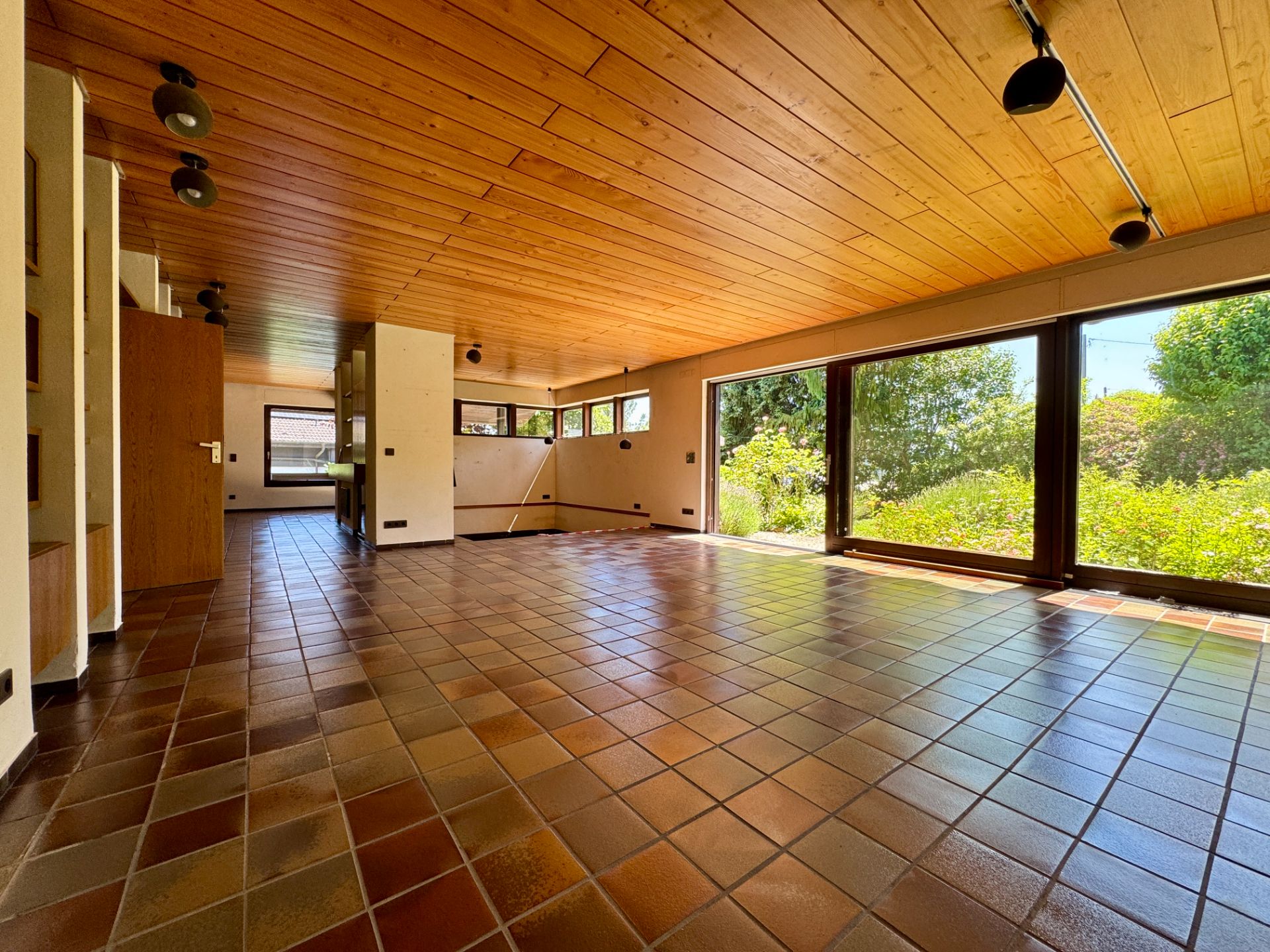 Großer, leerer Raum mit Fliesenboden, Holzdecke und großen Fenstern mit Blick auf Garten.