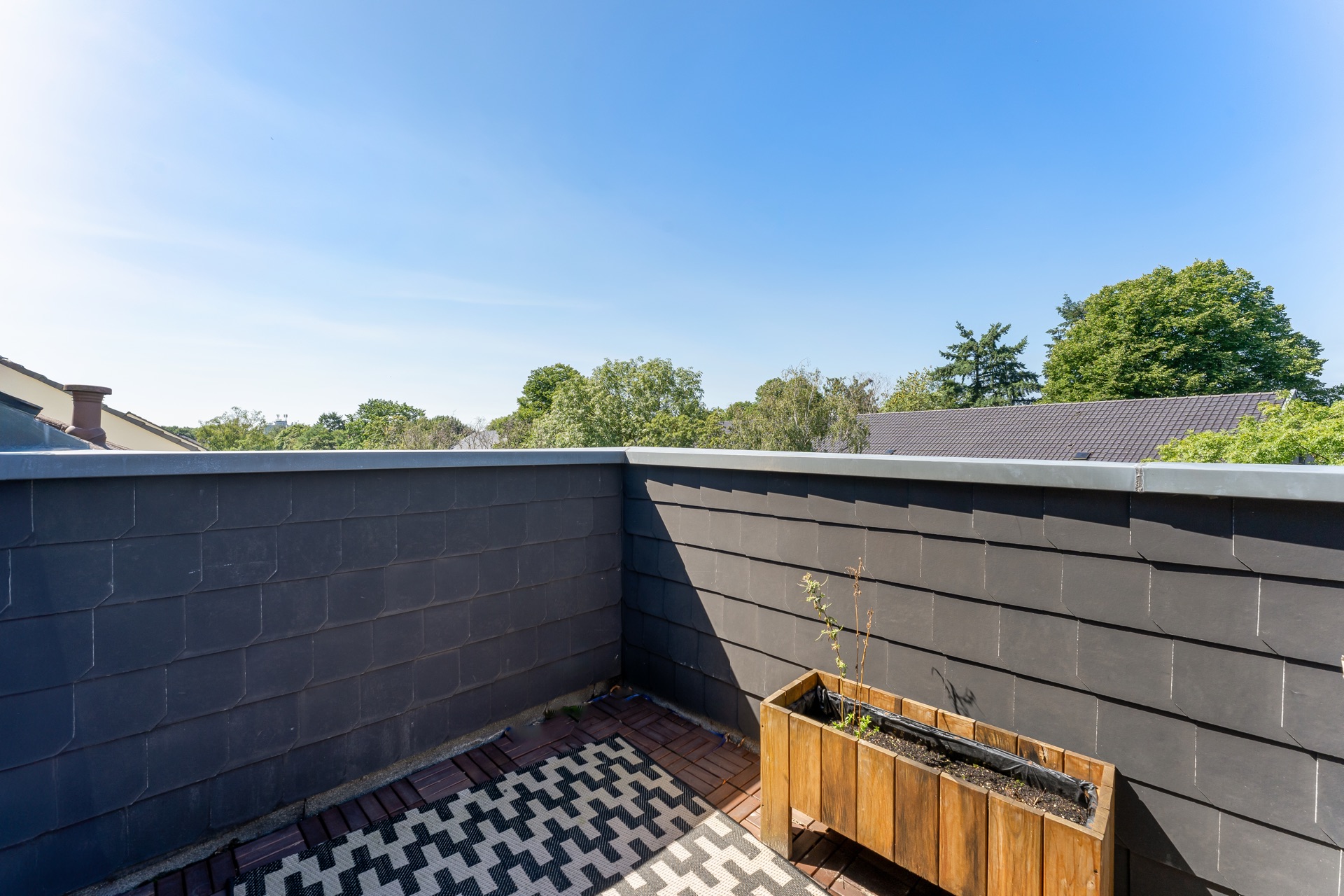 Kleine Dachterrasse mit dunkler Mauer, Holzkasten und Blick auf grüne Bäume.