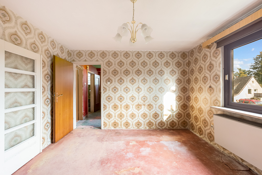 Leeres Zimmer mit gemusterter Tapete, offenem Fenster und Tür, abgenutzter Boden.