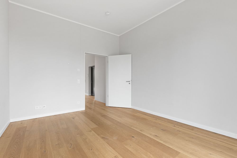 Leerer Raum mit hellem Holzboden, weißen Wänden und geöffneter weißer Tür.