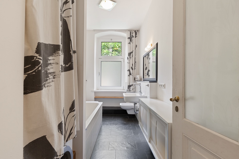 Helles Badezimmer mit Badewanne, Waschbecken, Spiegel und Fenster, schwarzer Fliesenboden.