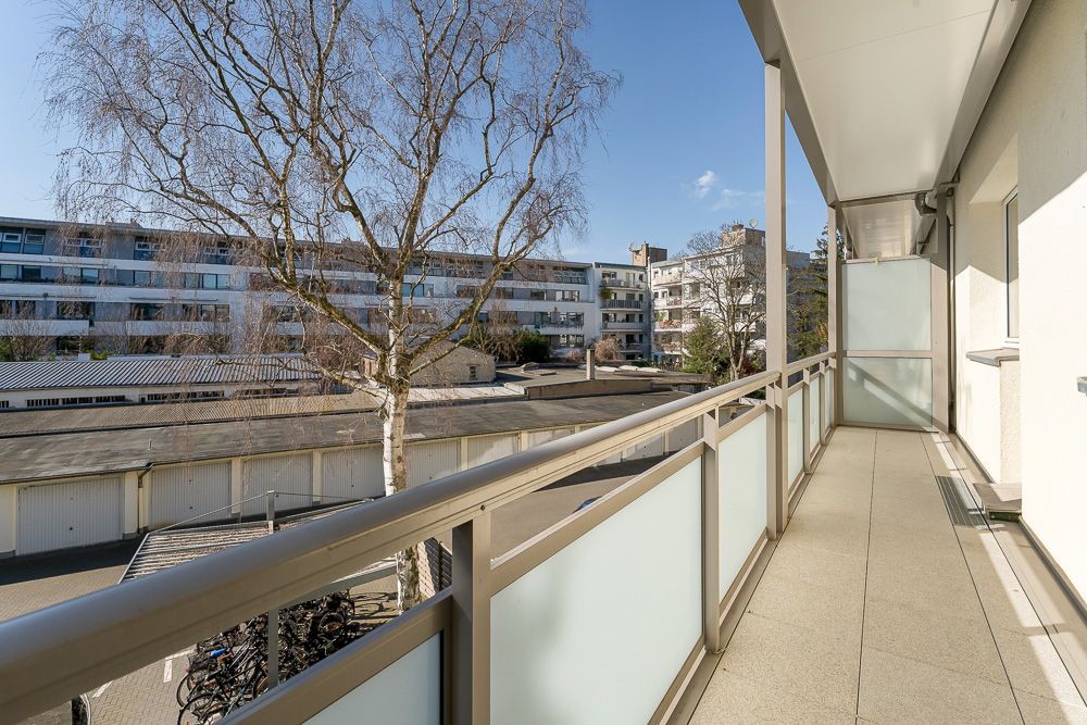 Langer Balkon mit Glasgeländer, Blick auf Wohngebäude und einen Baum im Hof.