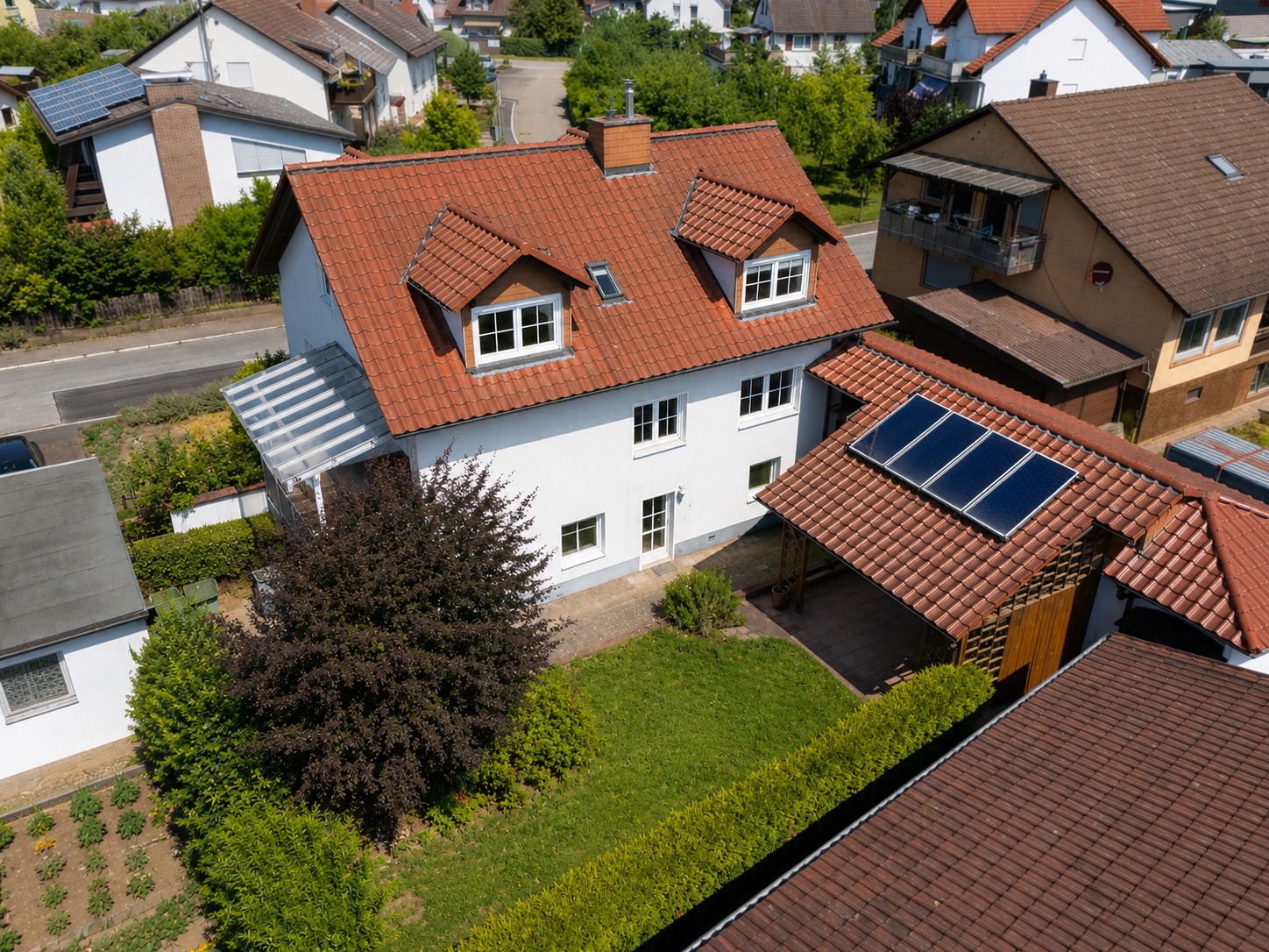 Weißes Wohnhaus mit rotem Dach, Garten und Carport mit Solarpanelen, aus der Vogelperspektive.