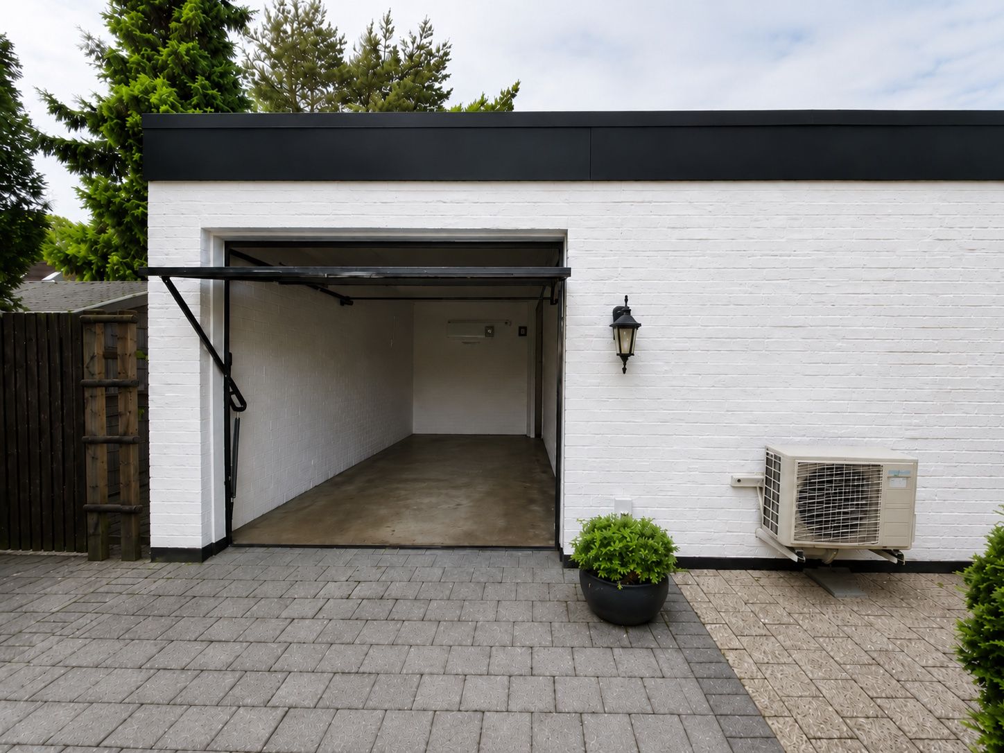Weiße Garage mit offenem Tor, daneben eine Wandlaterne, Klimagerät und ein Topf mit Pflanze.