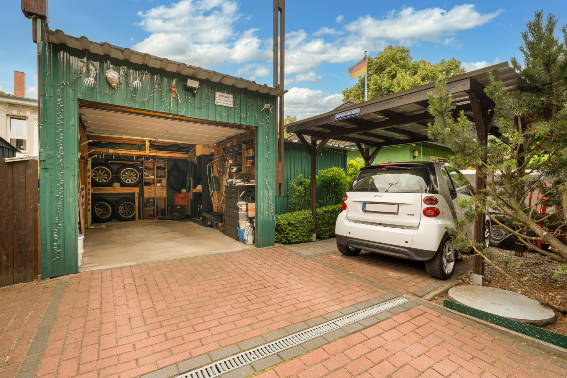 Eine offene Garage mit Werkzeugen und Reifen, daneben ein geparktes weißes Auto unter einem Carport.