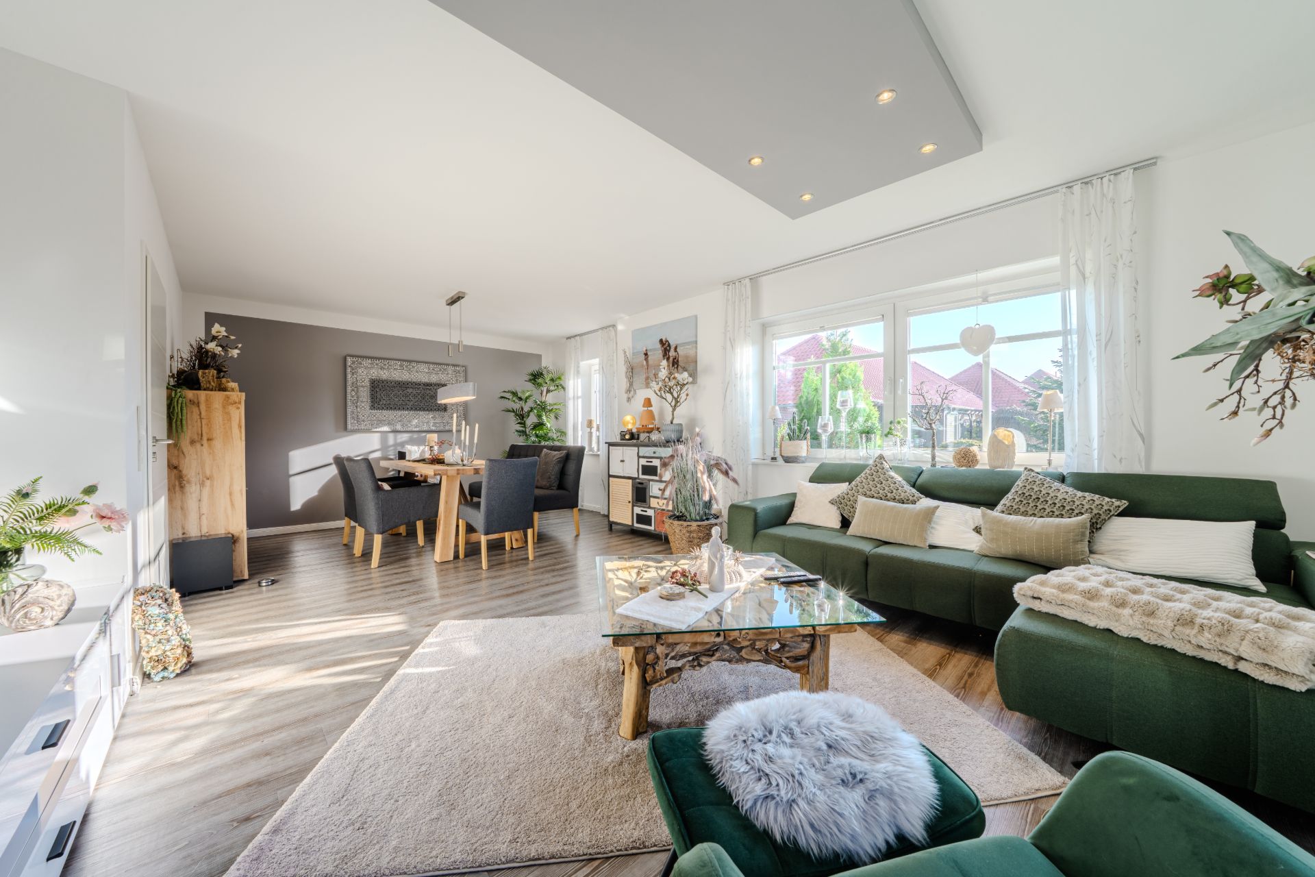 Helles Wohnzimmer mit grüner Couch, Glastisch, Essbereich und großen Fenstern.