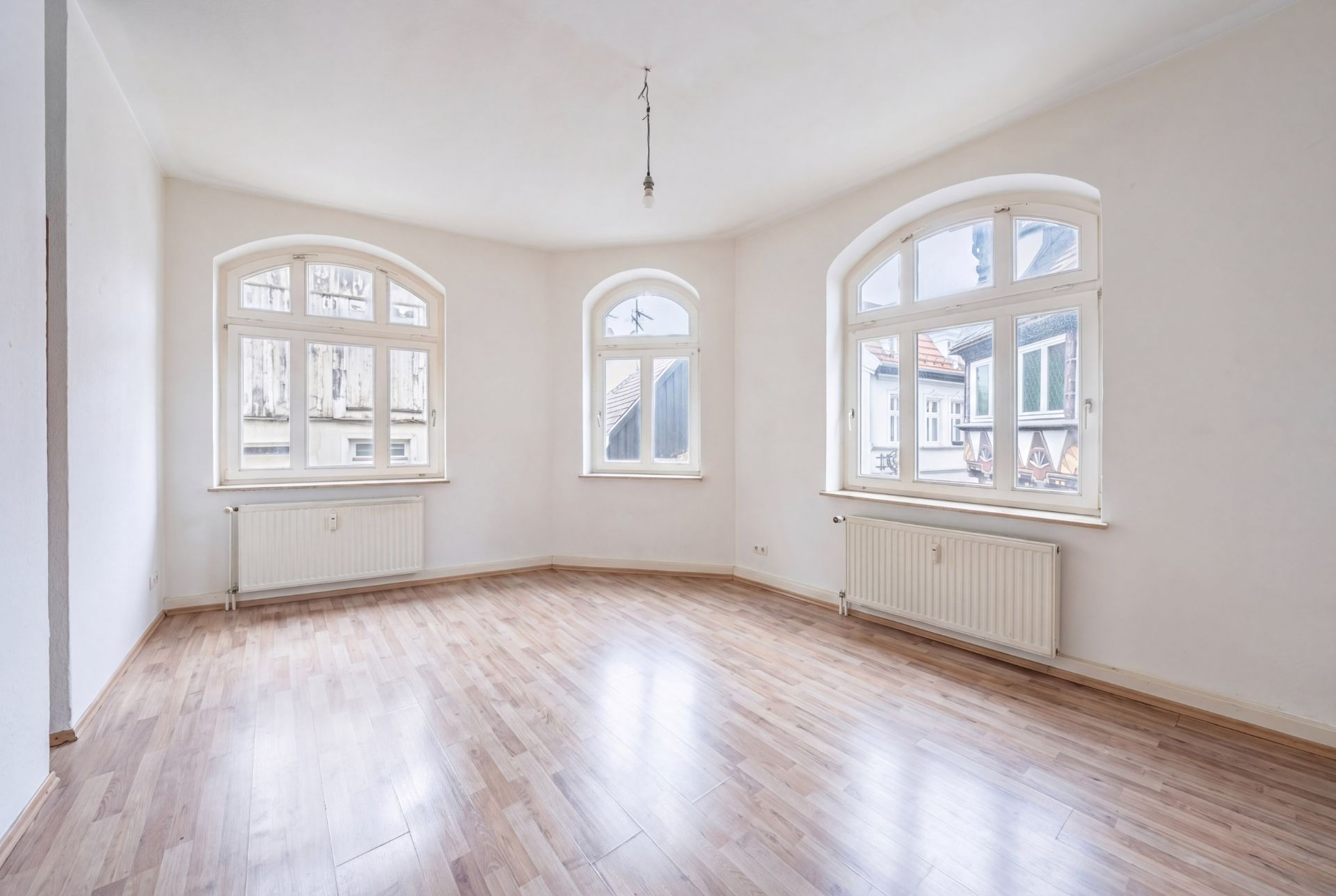 Leerer, heller Raum mit drei großen Fenstern und Holzfußboden.