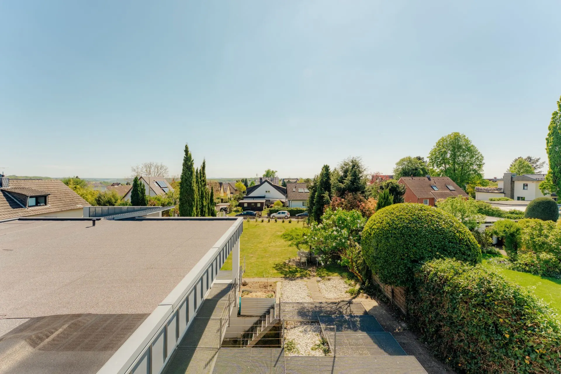 Blick auf einen Garten mit Rasen, Hecken und angrenzenden Wohnhäusern an einem sonnigen Tag.