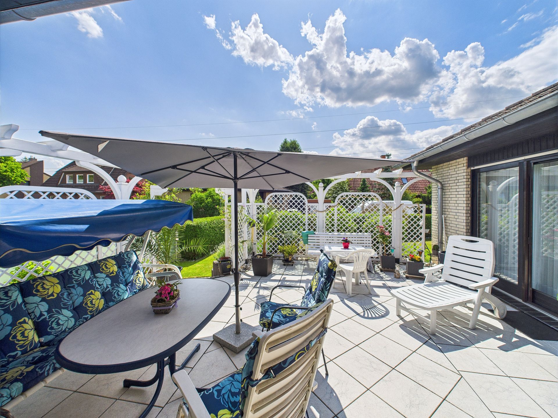 Sonnige Terrasse mit Gartenmöbeln, Sonnenschirm und Blumen, umgeben von weißen Gitterzäunen.