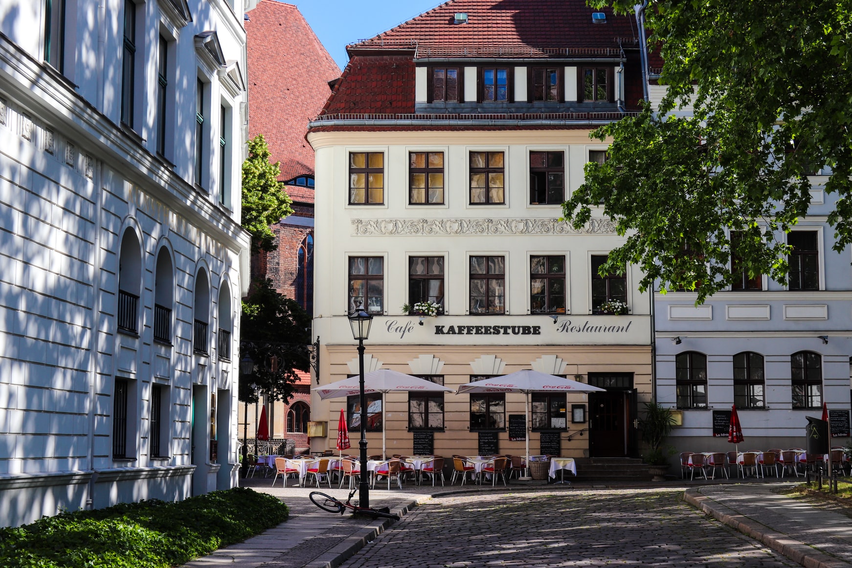 Straßenszene mit Café, Außenbestuhlung, Sonnenschirmen und historischen Gebäuden.