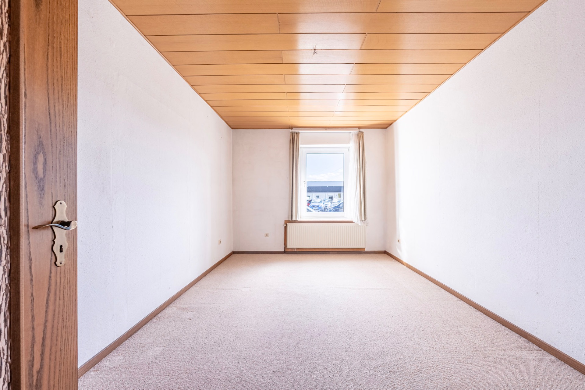 Helles, leeres Zimmer mit beigem Teppich, Holzdecke und Fenster am Ende.