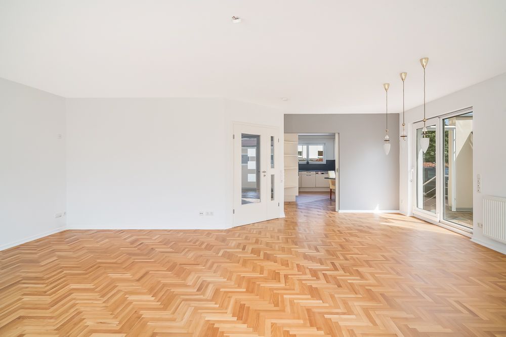 Helles Wohnzimmer mit Parkettboden, offenen Türen zur Küche und großen Fenstern zum Balkon.