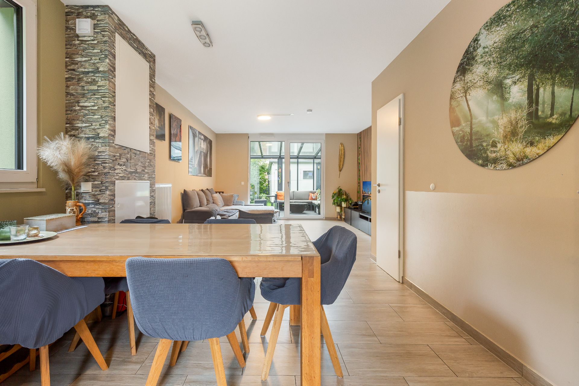 Essbereich mit Holztisch und blauen Stühlen, dahinter ein Wohnzimmer mit Sofa und Glastüren zum Garten.