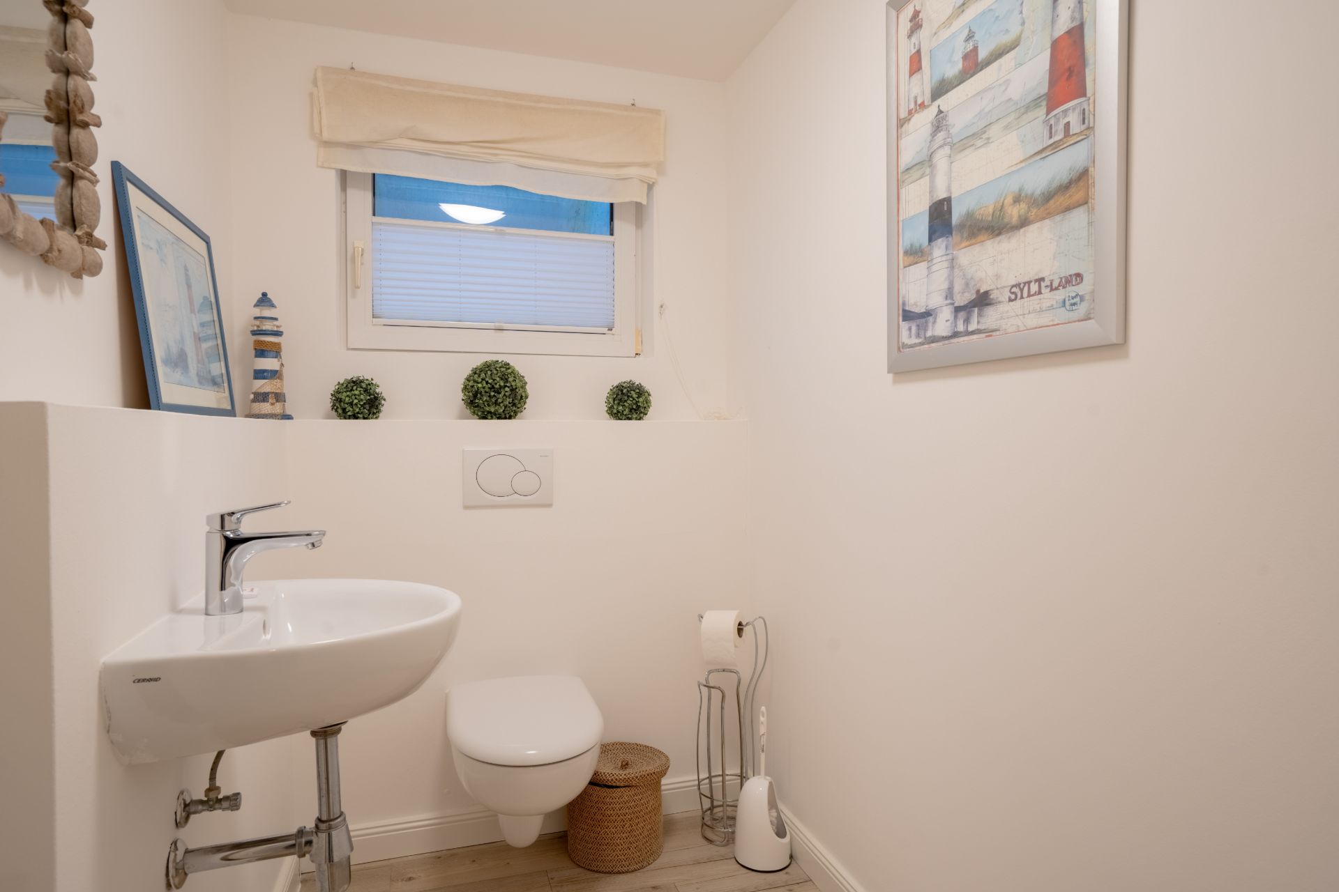 Helles Badezimmer mit Wandtoilette, Waschbecken, Fenster und maritimer Dekoration.