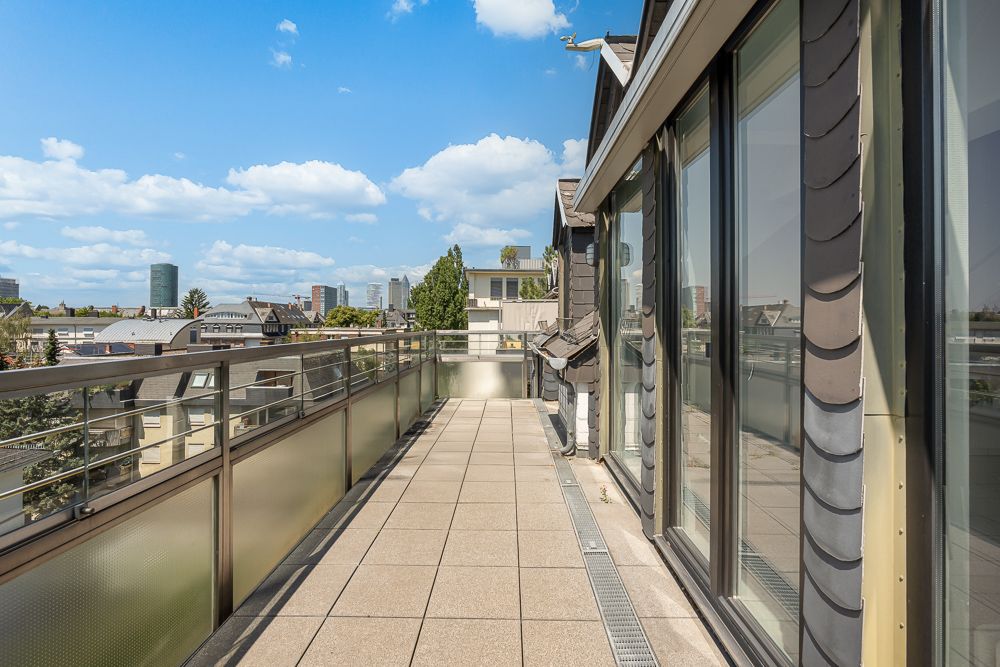 Großer Balkon mit Glasgeländer und Blick auf Stadtgebäude bei blauem Himmel.