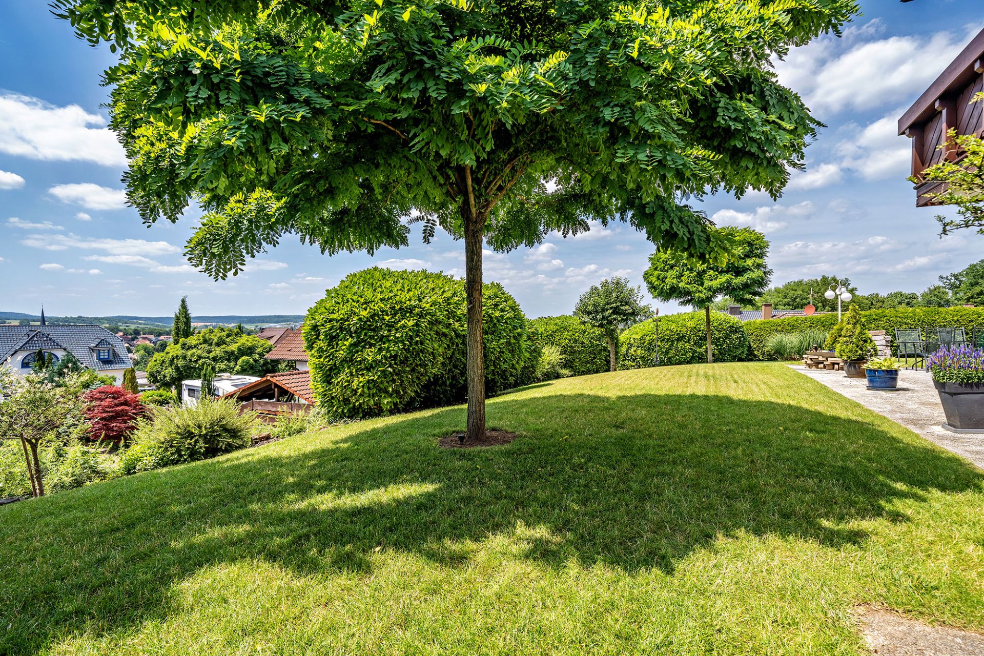 Grüner Garten mit Rasenfläche, Bäumen, Hecken und Terrasse mit Blumenkübeln.