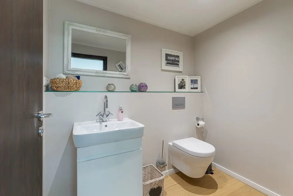 Modernes Badezimmer mit Wandtoilette, Waschbecken, Spiegel und Dekoration auf Glasablage.