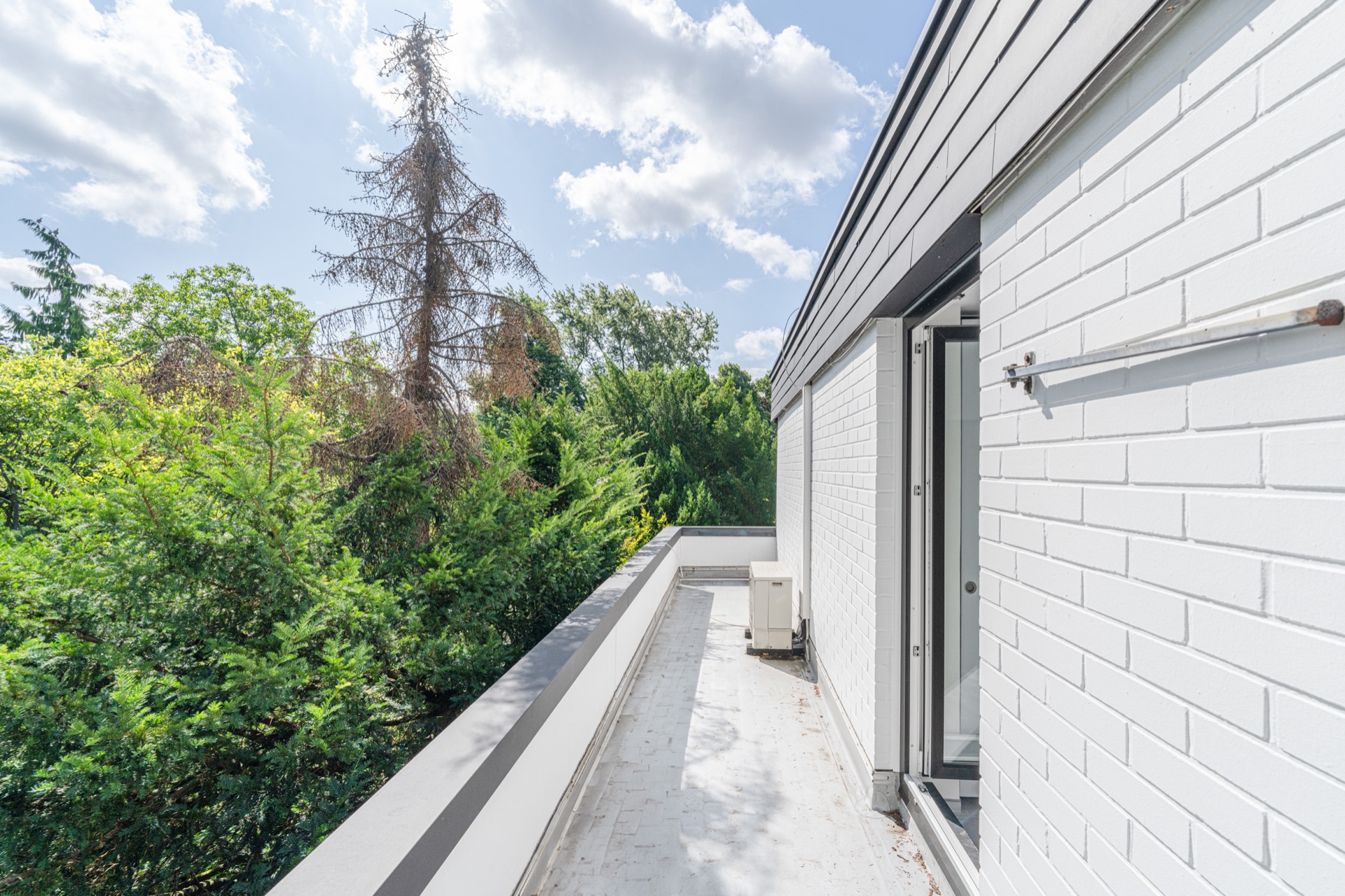 Helle Dachterrasse an einem weißen Gebäude mit Tür und Blick auf grüne Bäume.