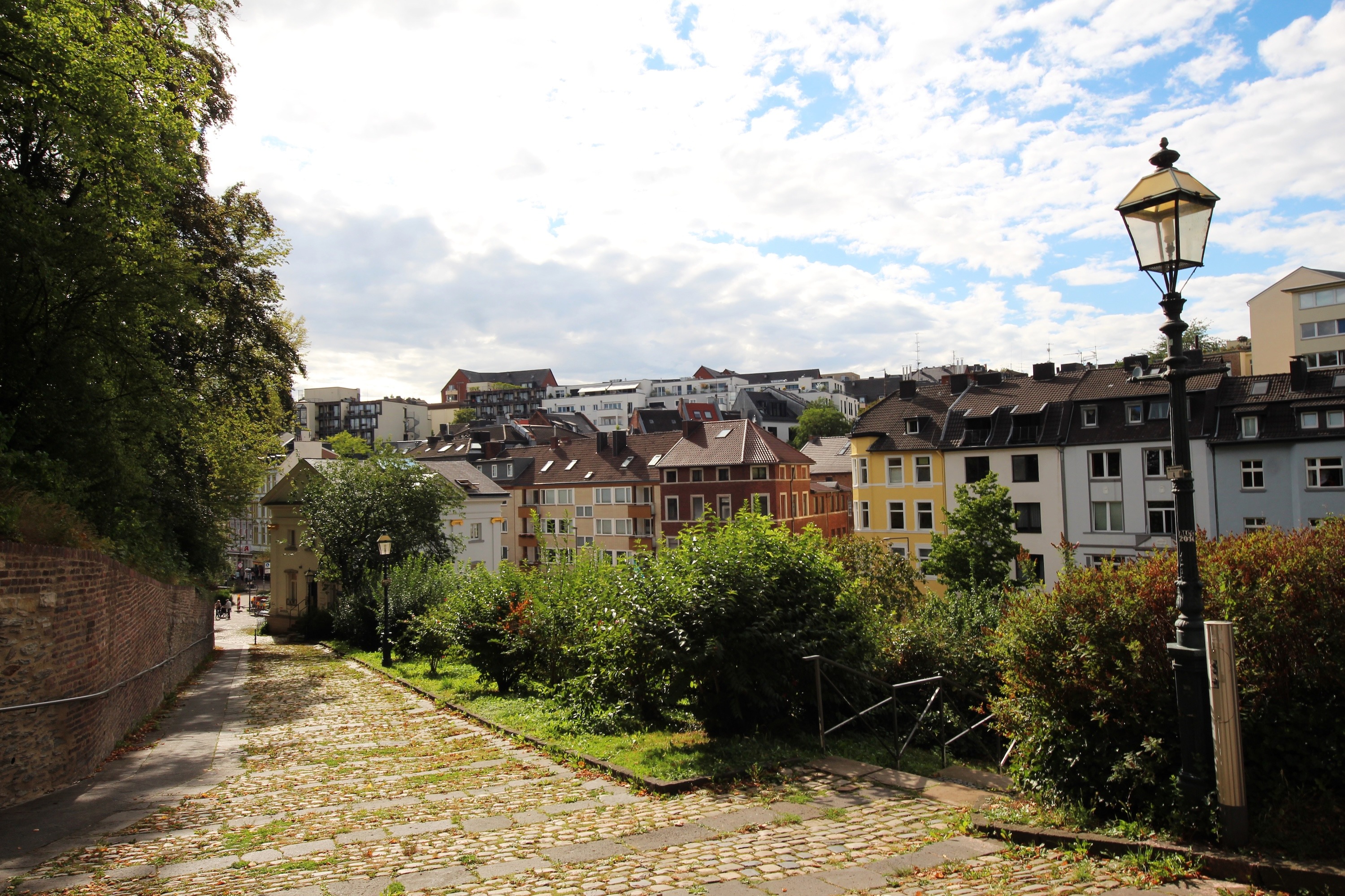 Gepflasterter Weg mit Laternen, Bäumen und Wohnhäusern im Hintergrund.