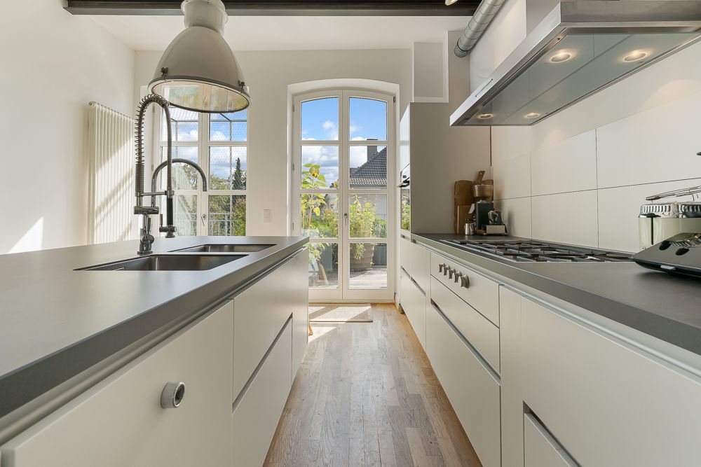 Moderne, schmale Küche mit weißen Schränken, Kochfeld, Spüle und großem Fenster zur Terrasse.