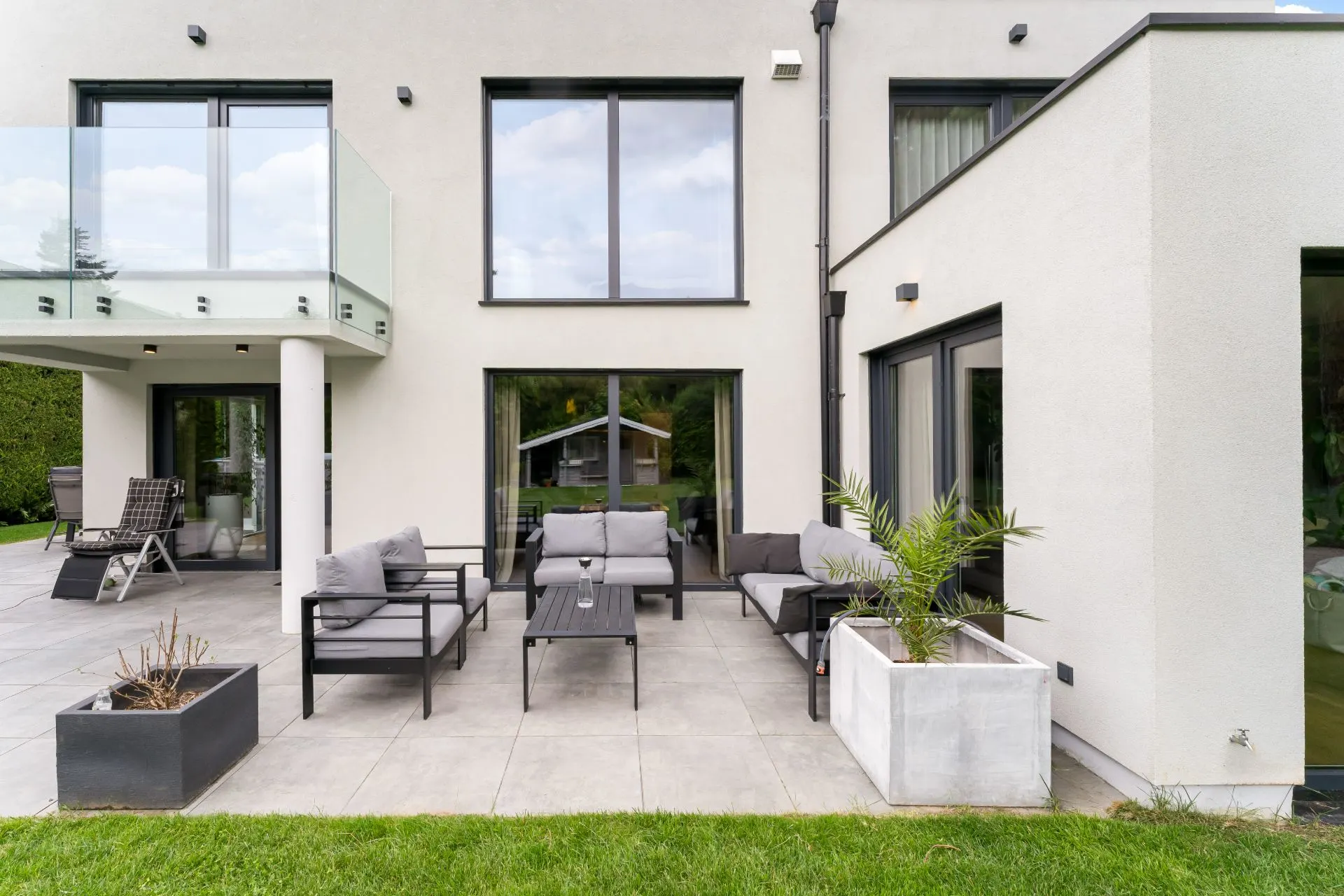 Moderne Terrasse mit Sofas, Pflanzenkübeln und großen Fenstern am Wohnhaus.