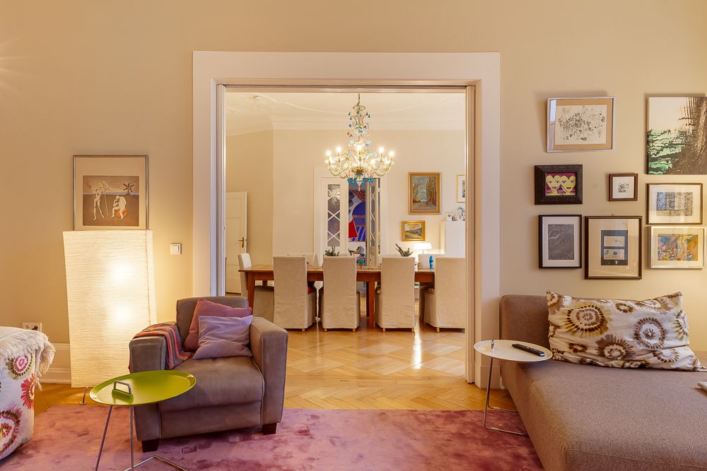 Helles Wohnzimmer mit Sessel, Sofa und Blick auf Esszimmer mit großem Tisch und Kronleuchter.