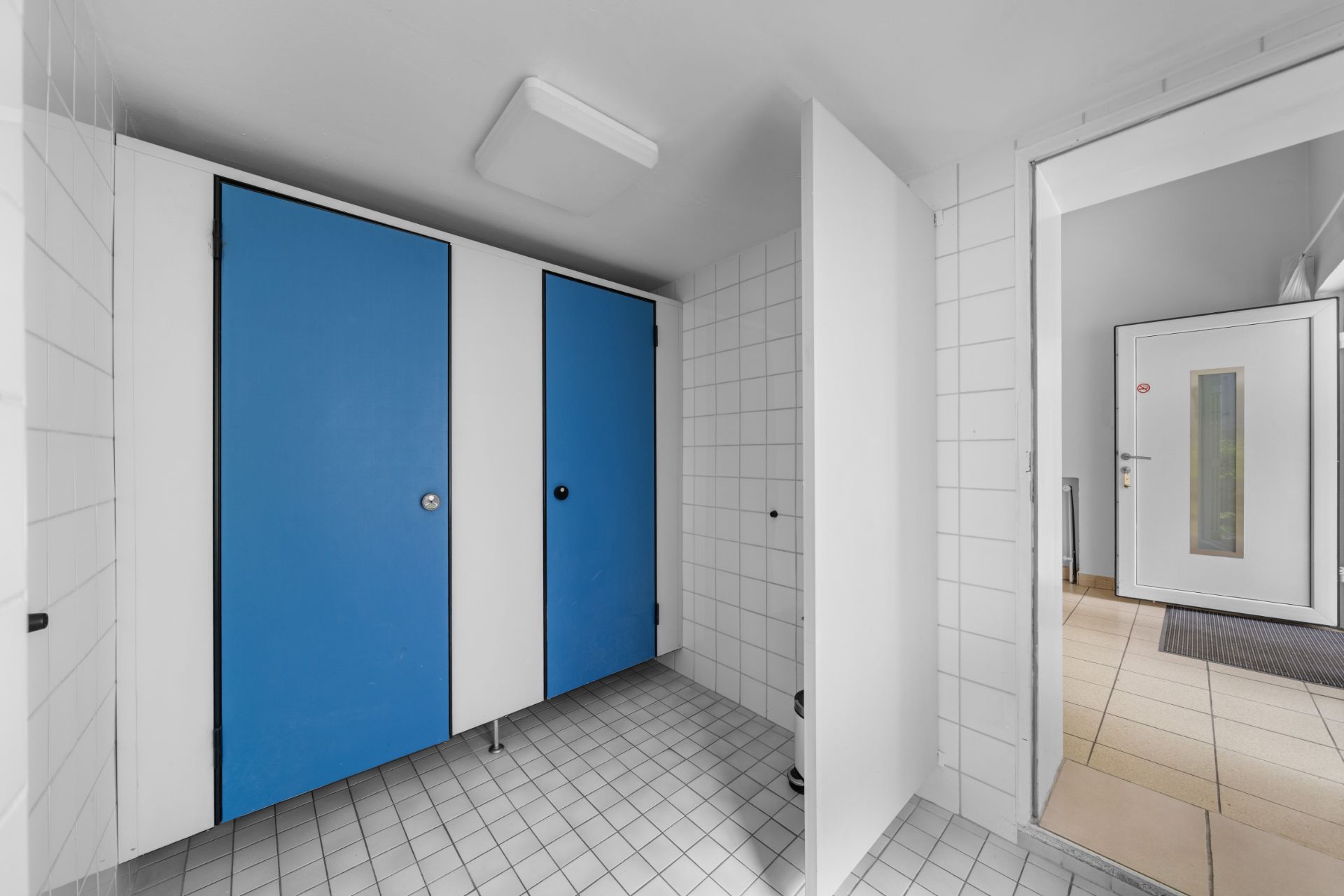 Weiß gefliester Raum mit zwei blauen Kabinentüren und einer offenen weißen Tür.