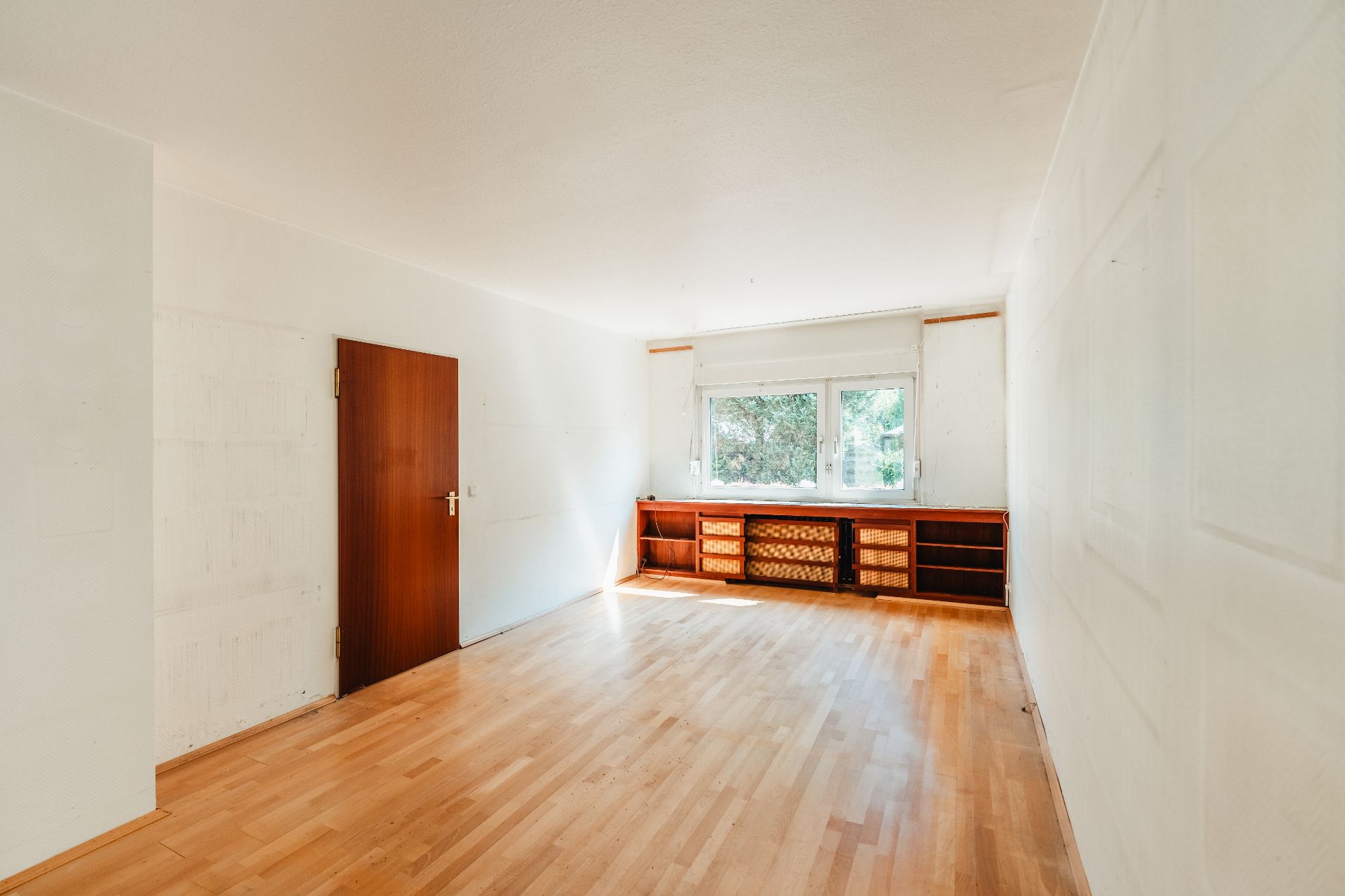 Helles, leeres Zimmer mit Holzboden, Holztür und großem Fenster mit Regalen darunter.