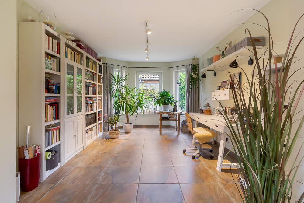 Helles Arbeitszimmer mit großen Fenstern, vielen Pflanzen, Bücherregal und Schreibtisch.