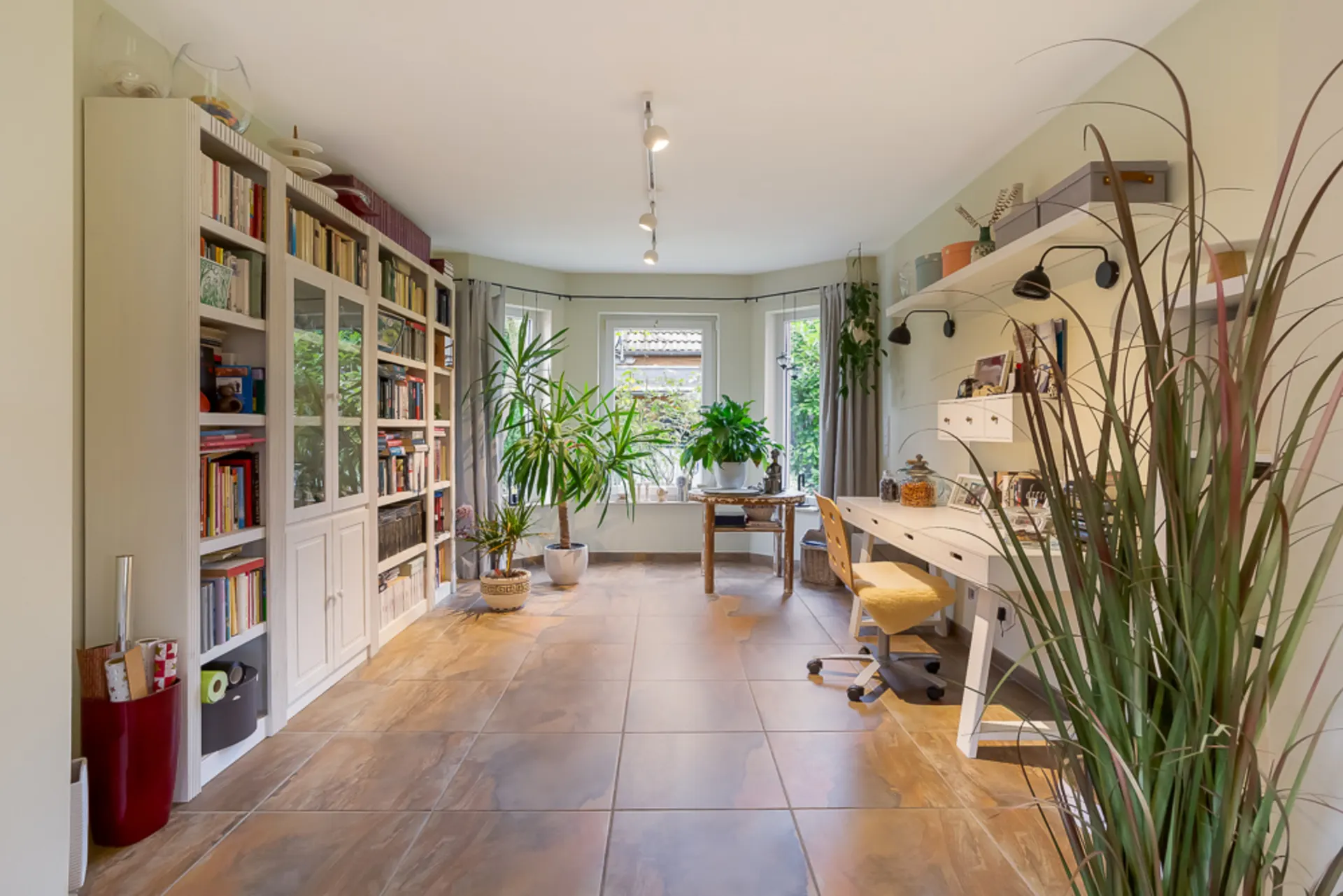 Helles Arbeitszimmer mit großen Fenstern, vielen Pflanzen, Bücherregal und Schreibtisch.