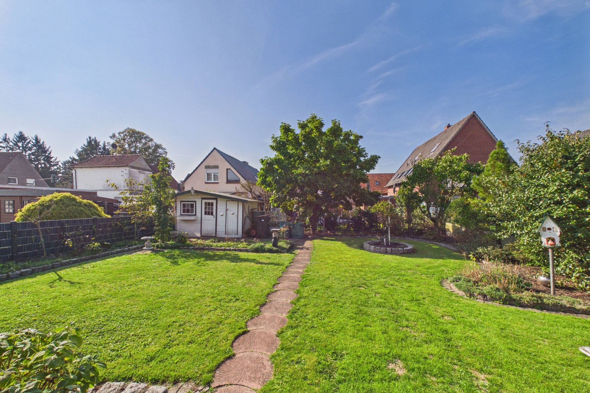 Garten mit Rasen, Steingehweg, Bäumen und einem kleinen Haus im Hintergrund.