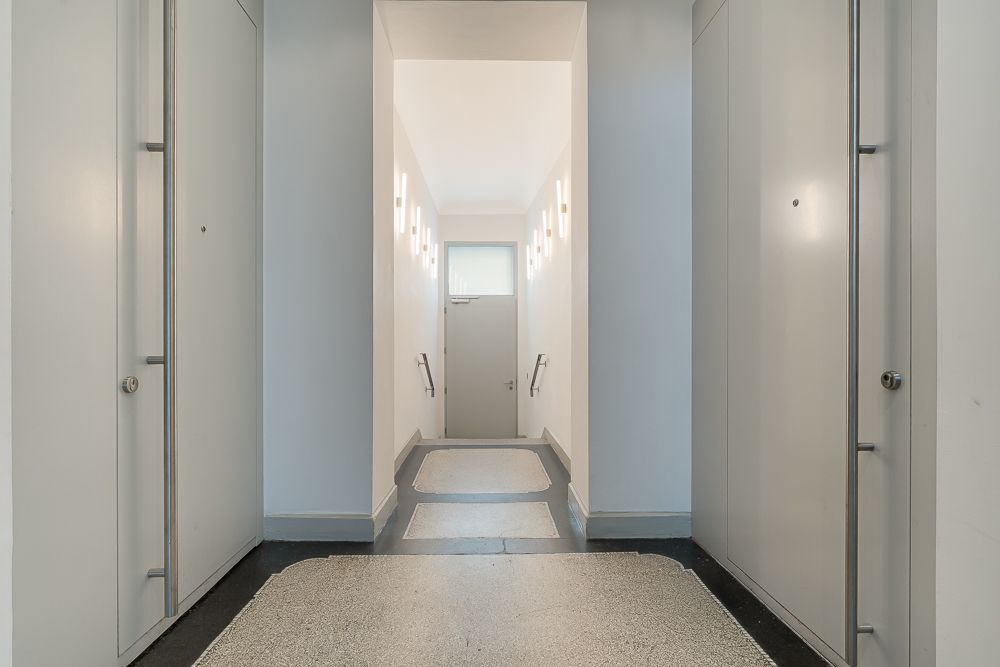Heller Flur mit grauer Tür, seitlichen Wandleuchten und Handläufen an den Wänden.