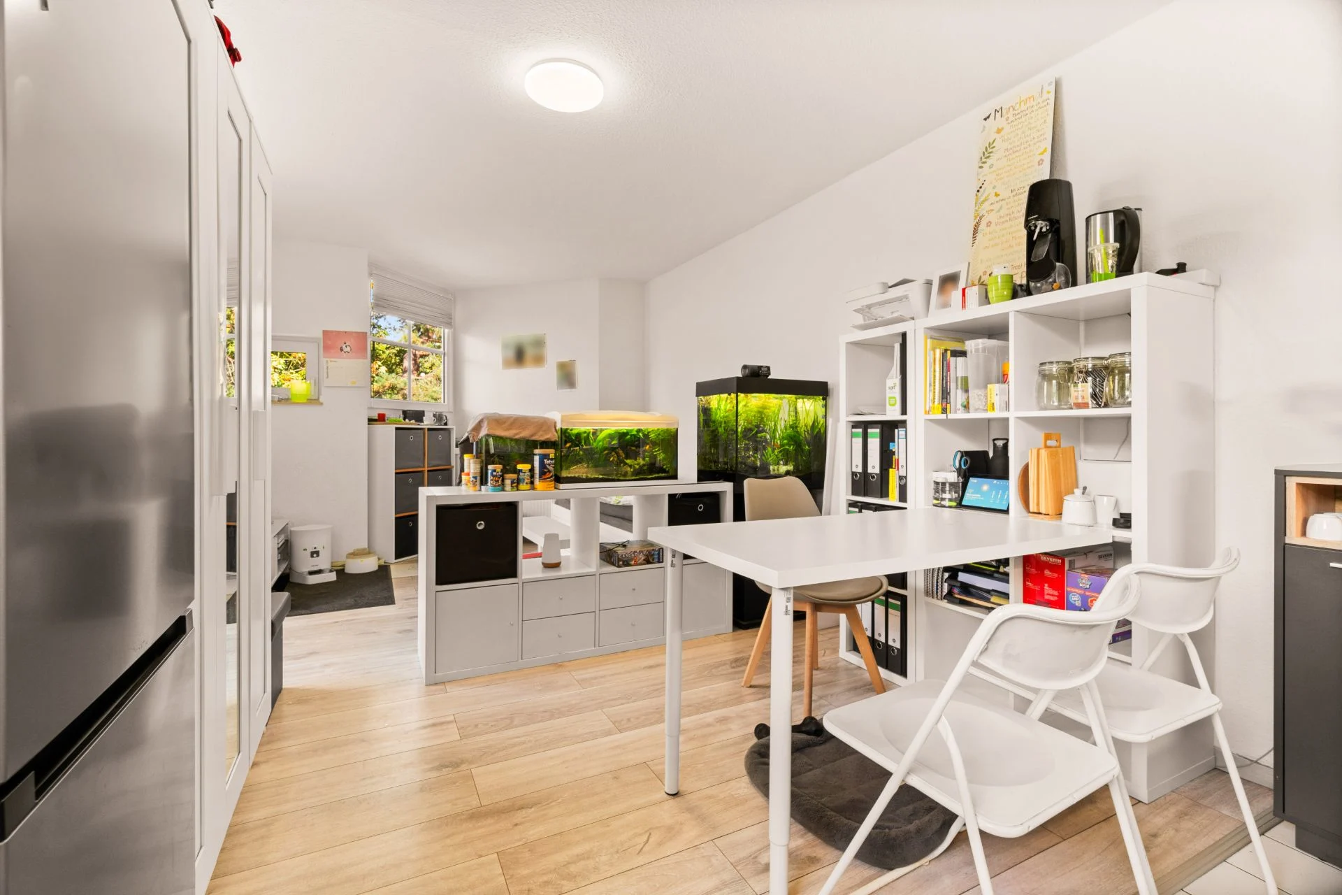 Helles Zimmer mit Tisch, zwei weißen Stühlen, Regalen und zwei Aquarien auf einem Sideboard.