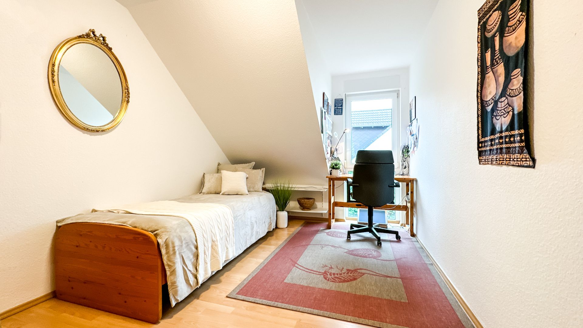 Helles Zimmer mit Einzelbett, Schreibtisch am Fenster, Teppich und Wandspiegel.