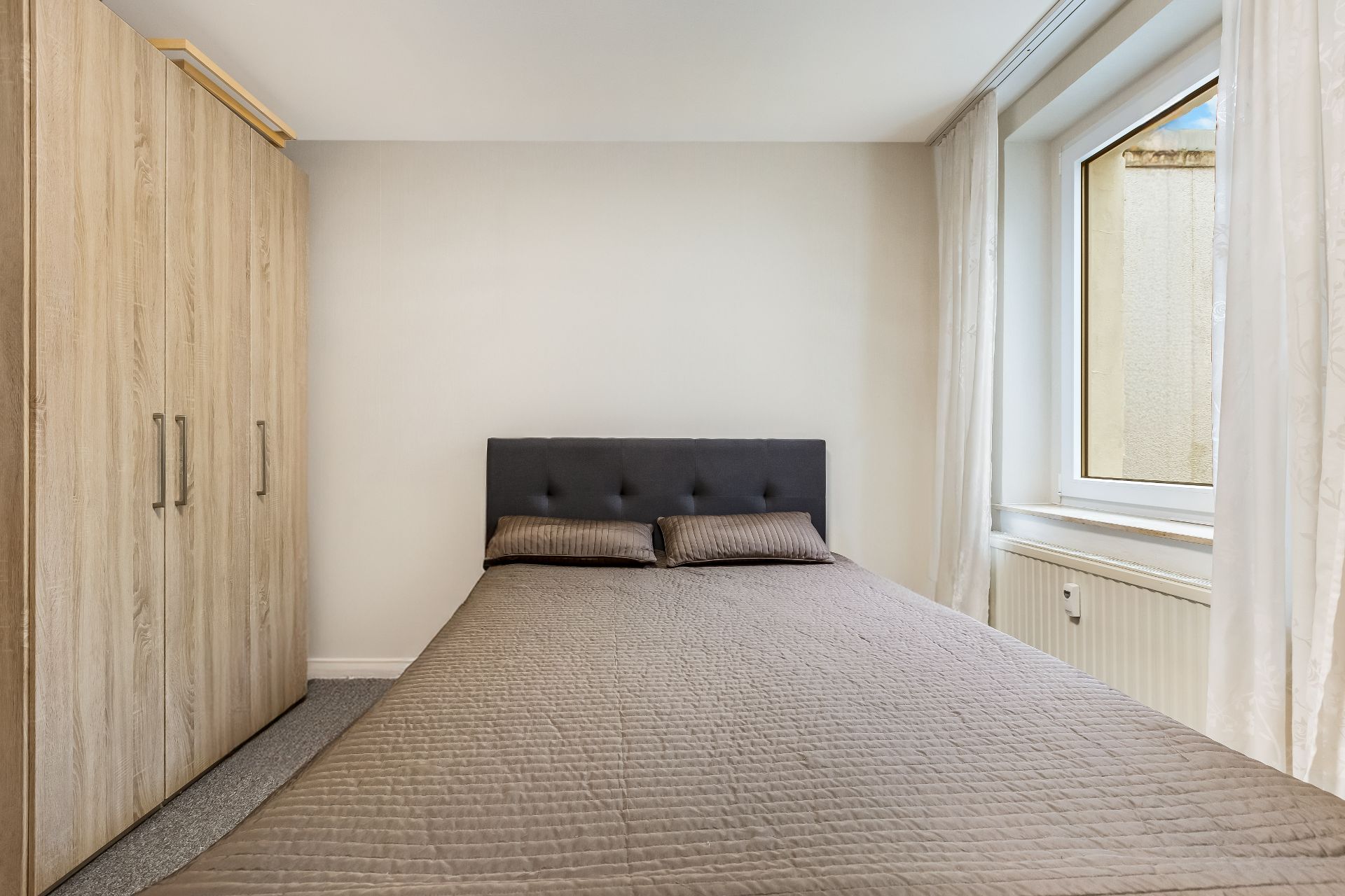 Schlafzimmer mit Doppelbett, zwei Kissen, Kleiderschrank und Fenster mit weißen Vorhängen.
