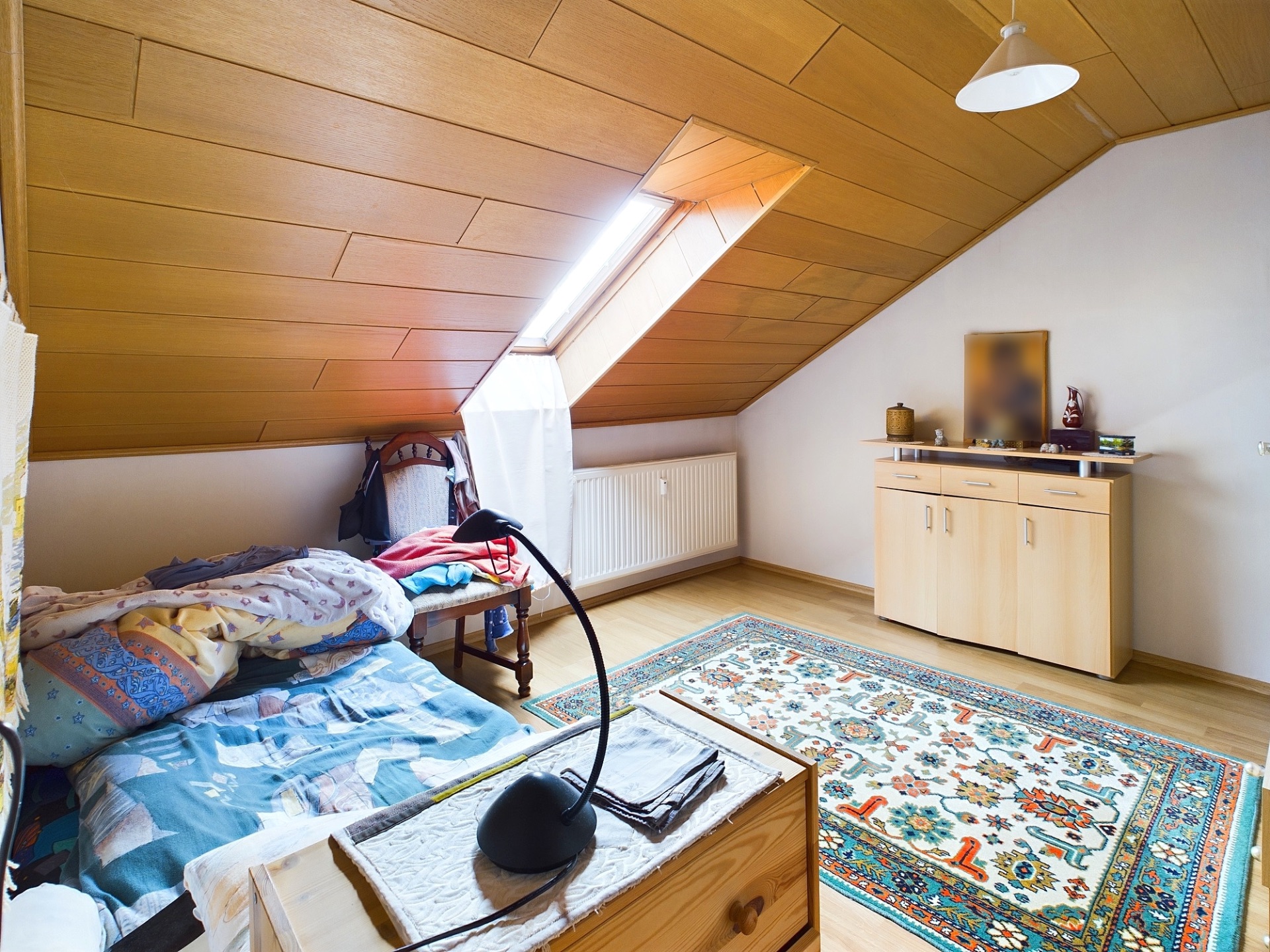 Dachzimmer mit Einzelbett, Teppich, Kommode und Schreibtischlampe unter schrägem Holzdachfenster.