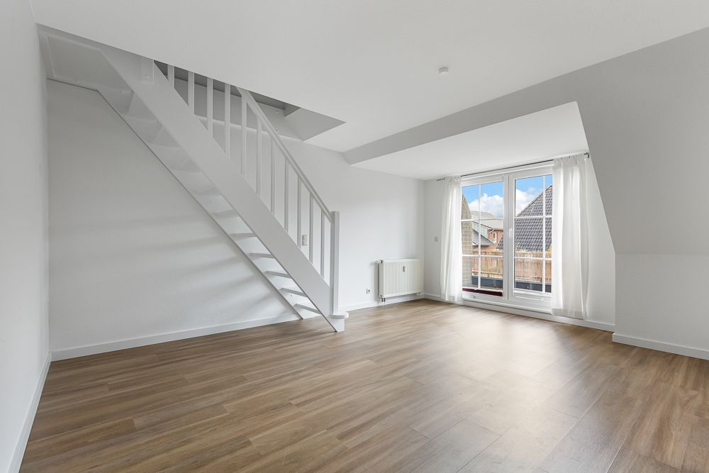 Heller, leerer Raum mit Holzfußboden, weißer Treppe und großem Fenster mit Blick nach draußen.