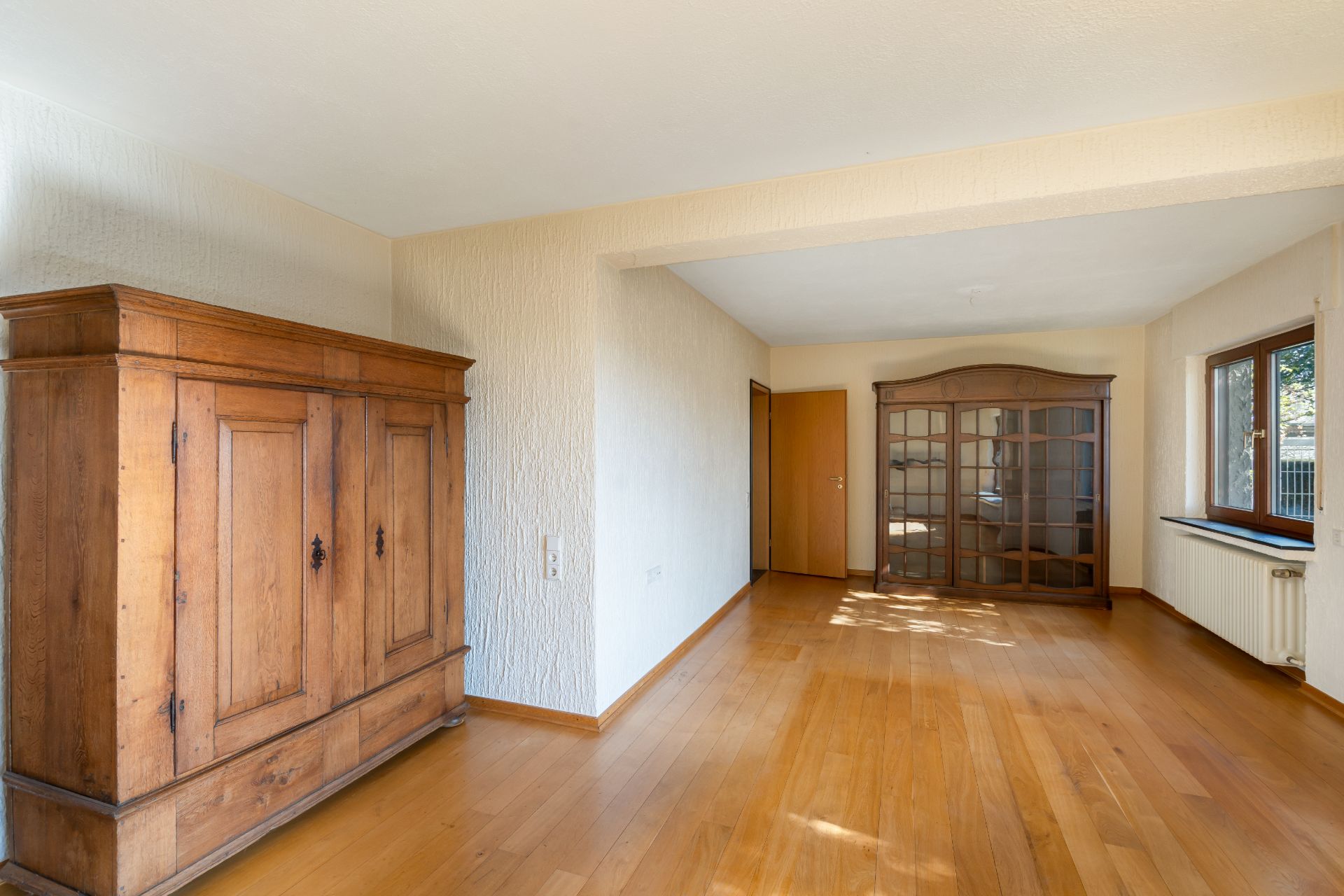 Helles Zimmer mit Holzboden, großem Holzschrank und Vitrine, Fenster rechts.