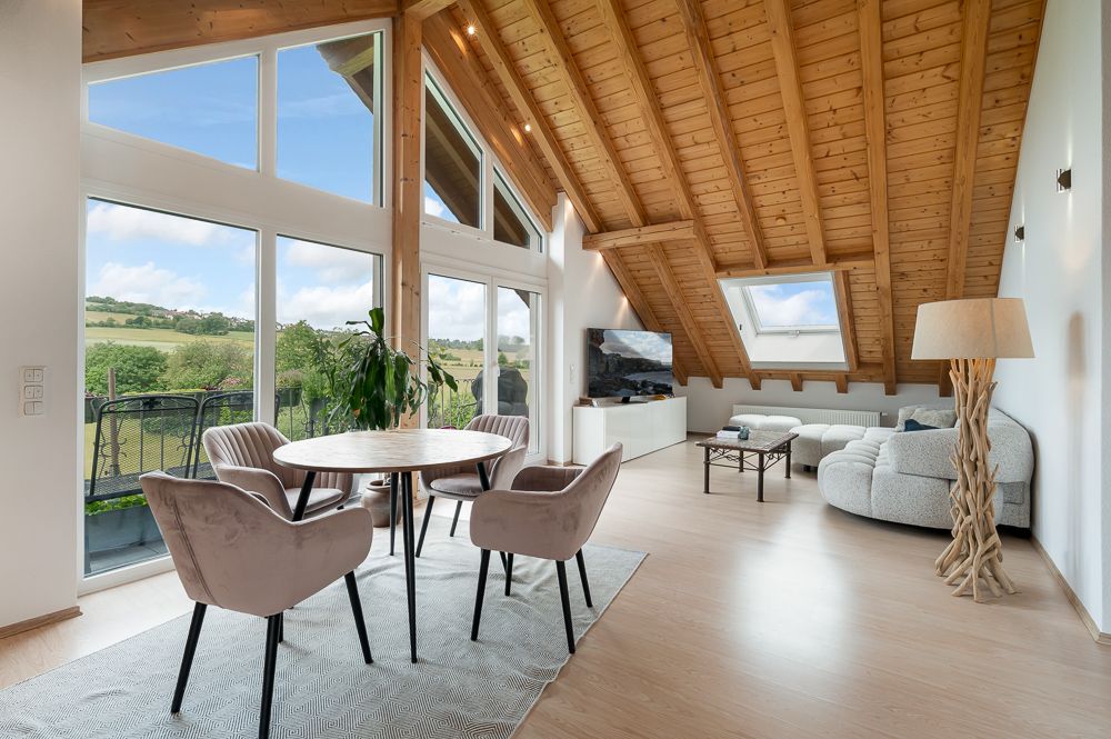 Helles Wohnzimmer mit Holzdach, Essbereich, großen Fenstern und Sitzgruppe.