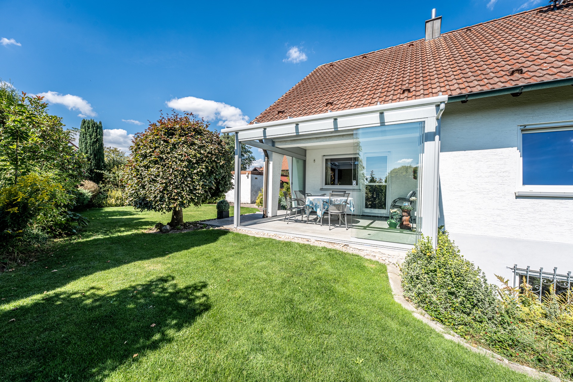 Garten mit überdachter Terrasse, Tisch und Stühlen an einem Wohnhaus bei sonnigem Wetter.