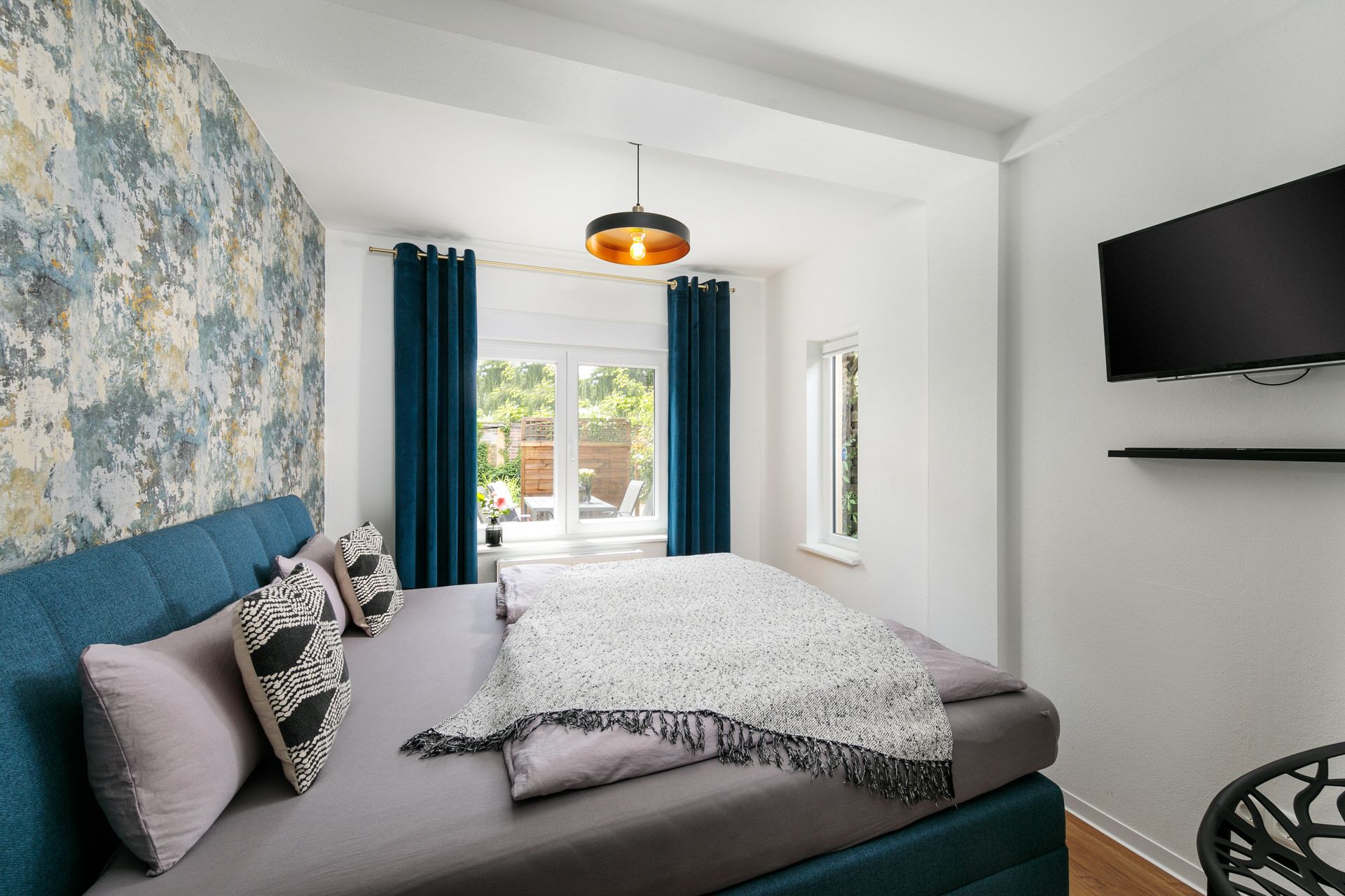 Helles Schlafzimmer mit Doppelbett, TV an der Wand und Fenster mit blauen Vorhängen.