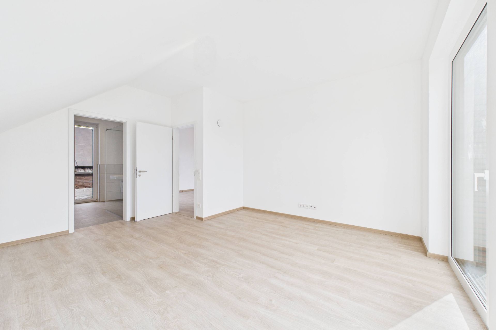 Helles, leeres Zimmer mit Holzboden, weißen Wänden und großem Fenster.