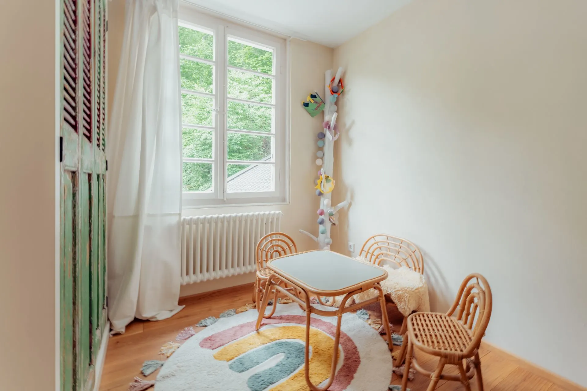 Helles Zimmer mit kleinem Tisch, zwei Stühlen und buntem Teppich vor einem Fenster.