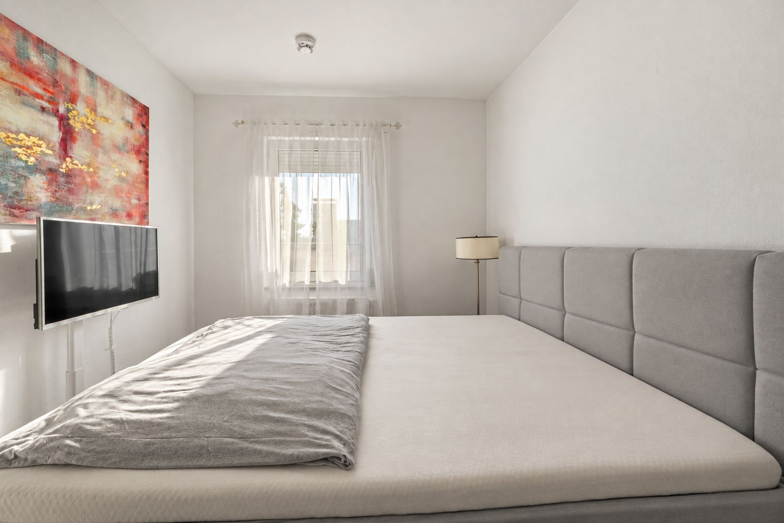 Helles Schlafzimmer mit grauem Bett, Wandbild, Fernseher und Fenster mit Gardine.