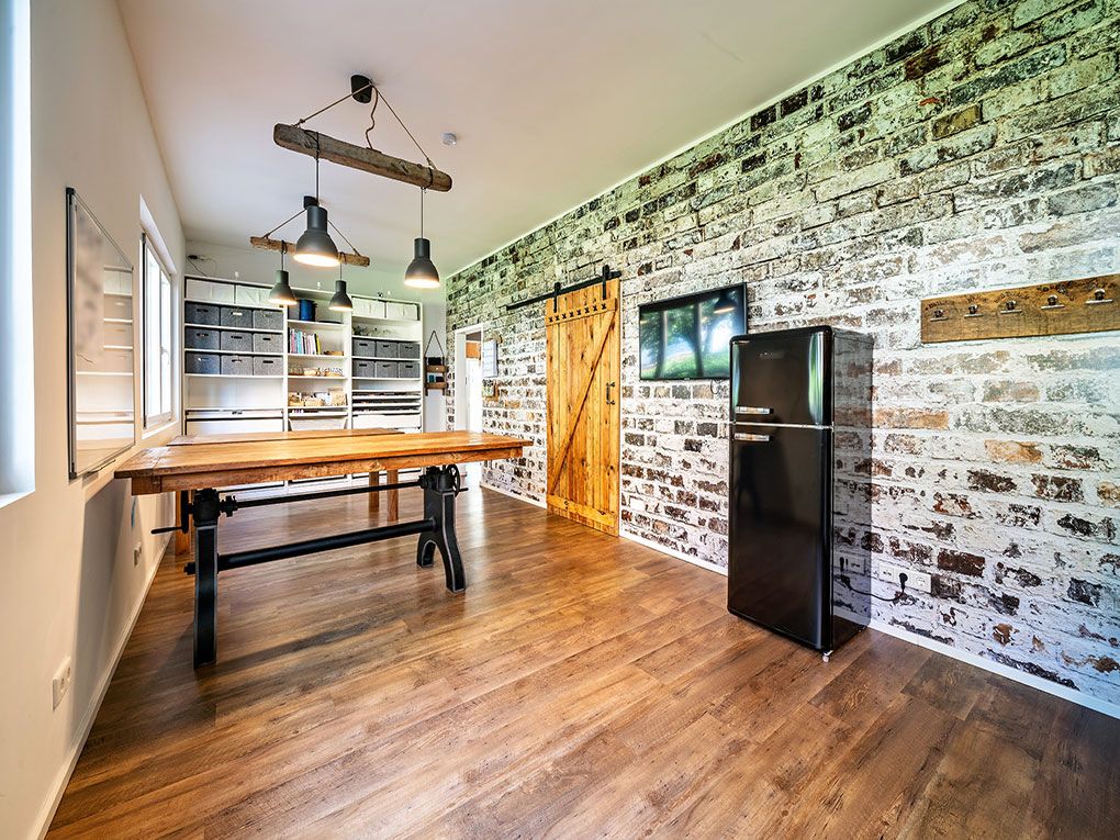 Raum mit Holztisch, schwarzen Kühlschrank, Regalwand und Ziegelwand mit Holztür.
