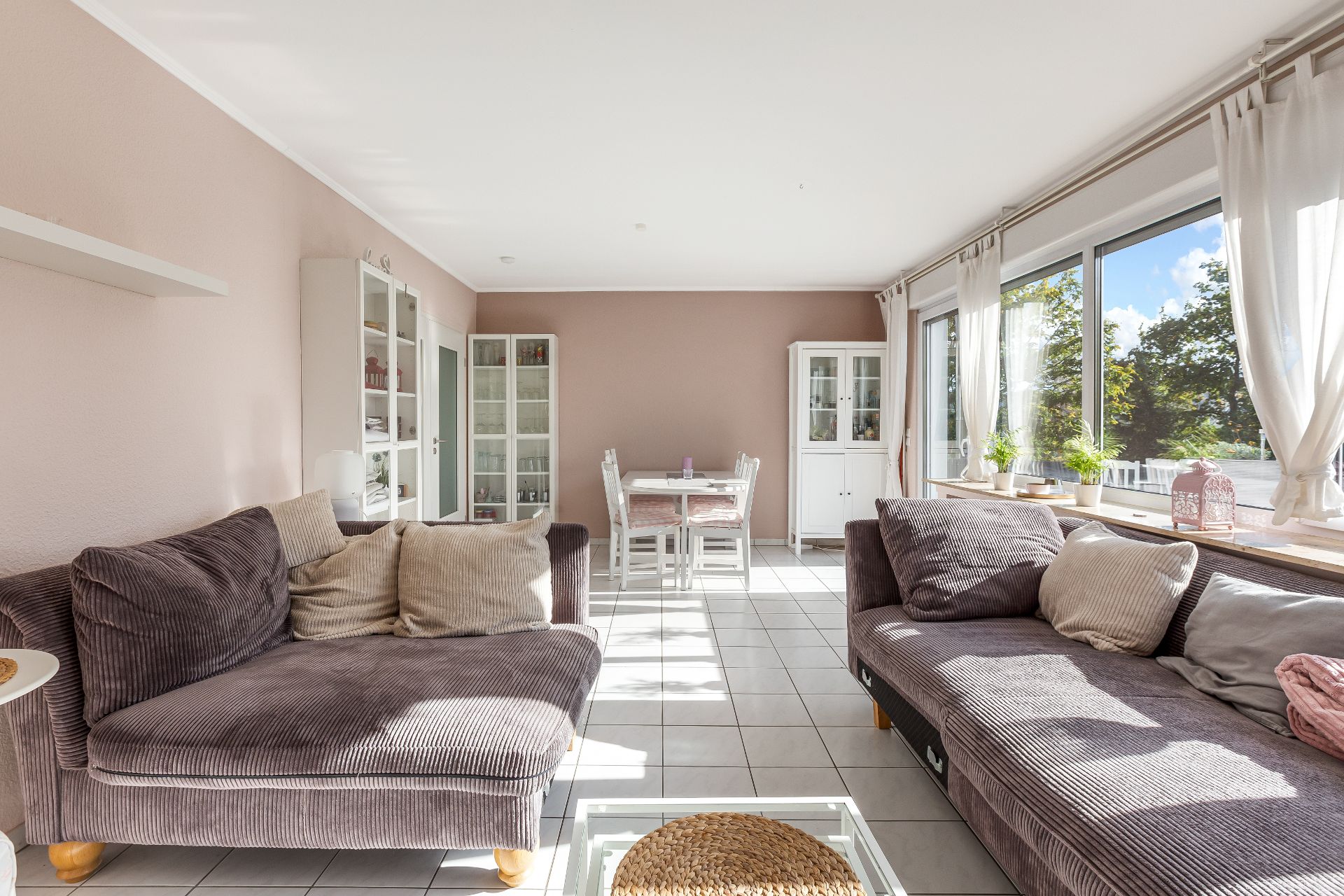 Helles Wohnzimmer mit zwei Sofas, Esstisch, Vitrinen und großen Fenstern mit Vorhängen.